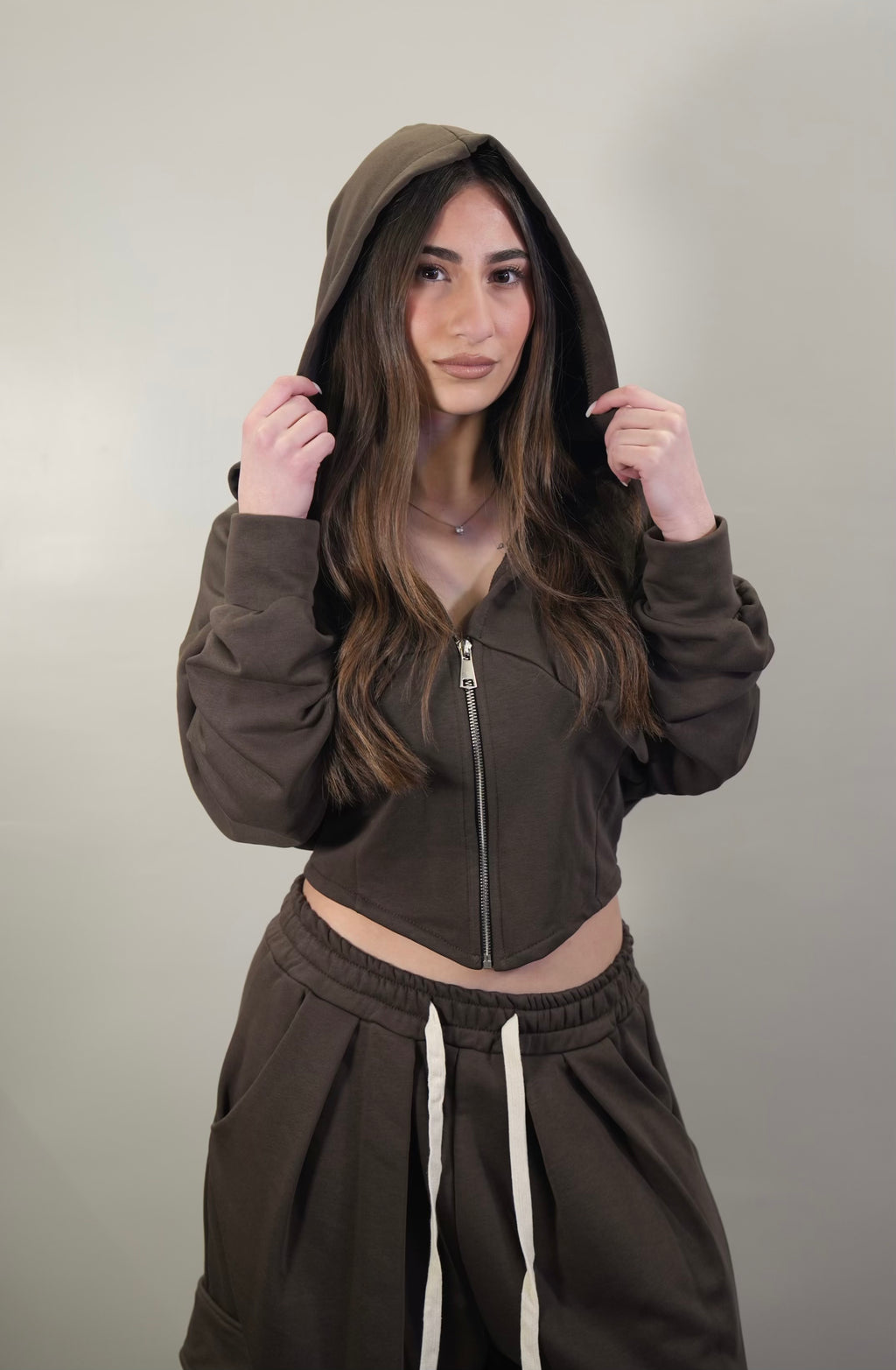 Primo piano della felpa crop marrone con zip aperta, cappuccio indossato e focus sul taglio moderno del capo.