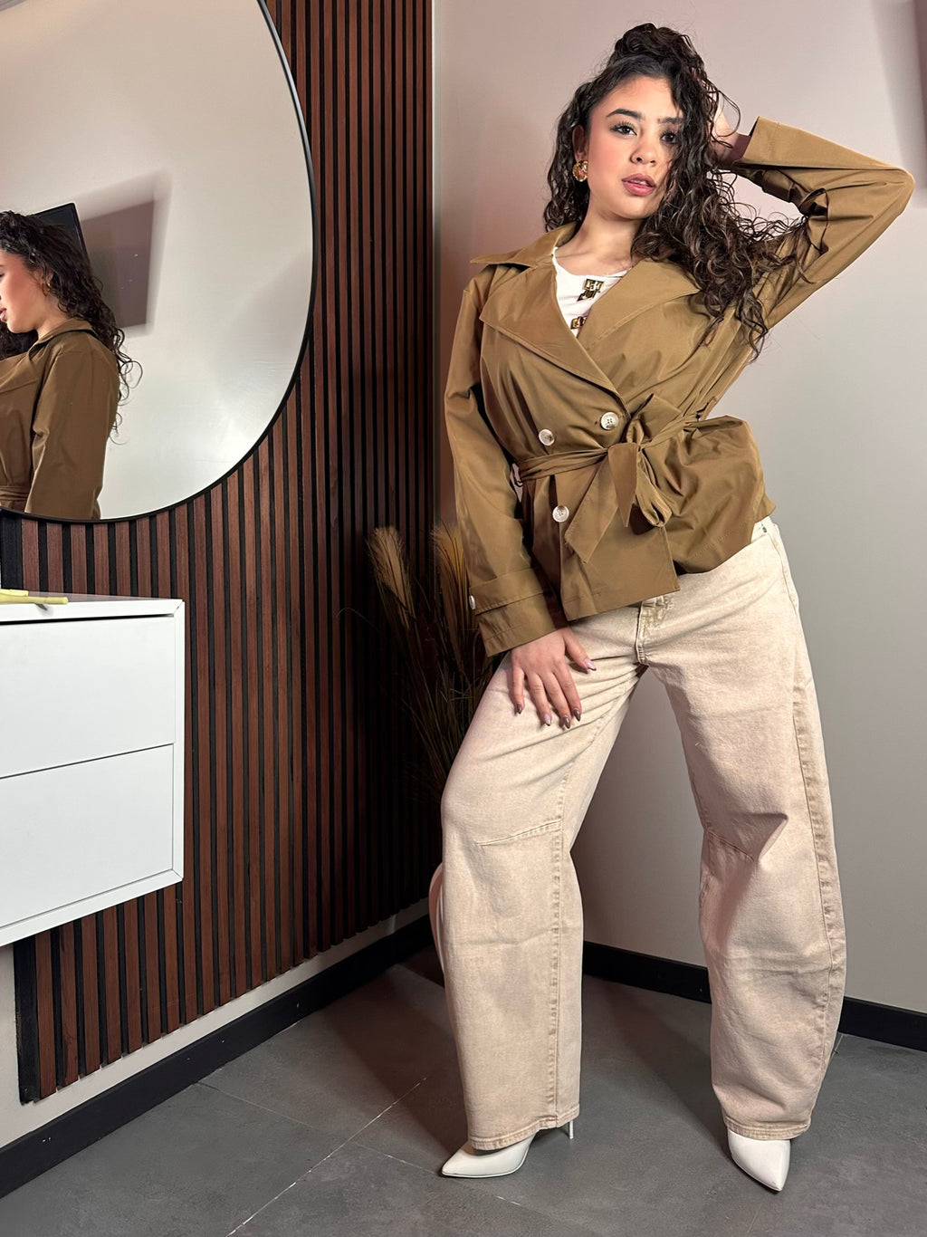 Posa a figura intera della modella con indosso il trench corto color cammello allacciato e pantaloni a zampa color panna. Lo sfondo mostra un pannello decorativo in legno scuro.