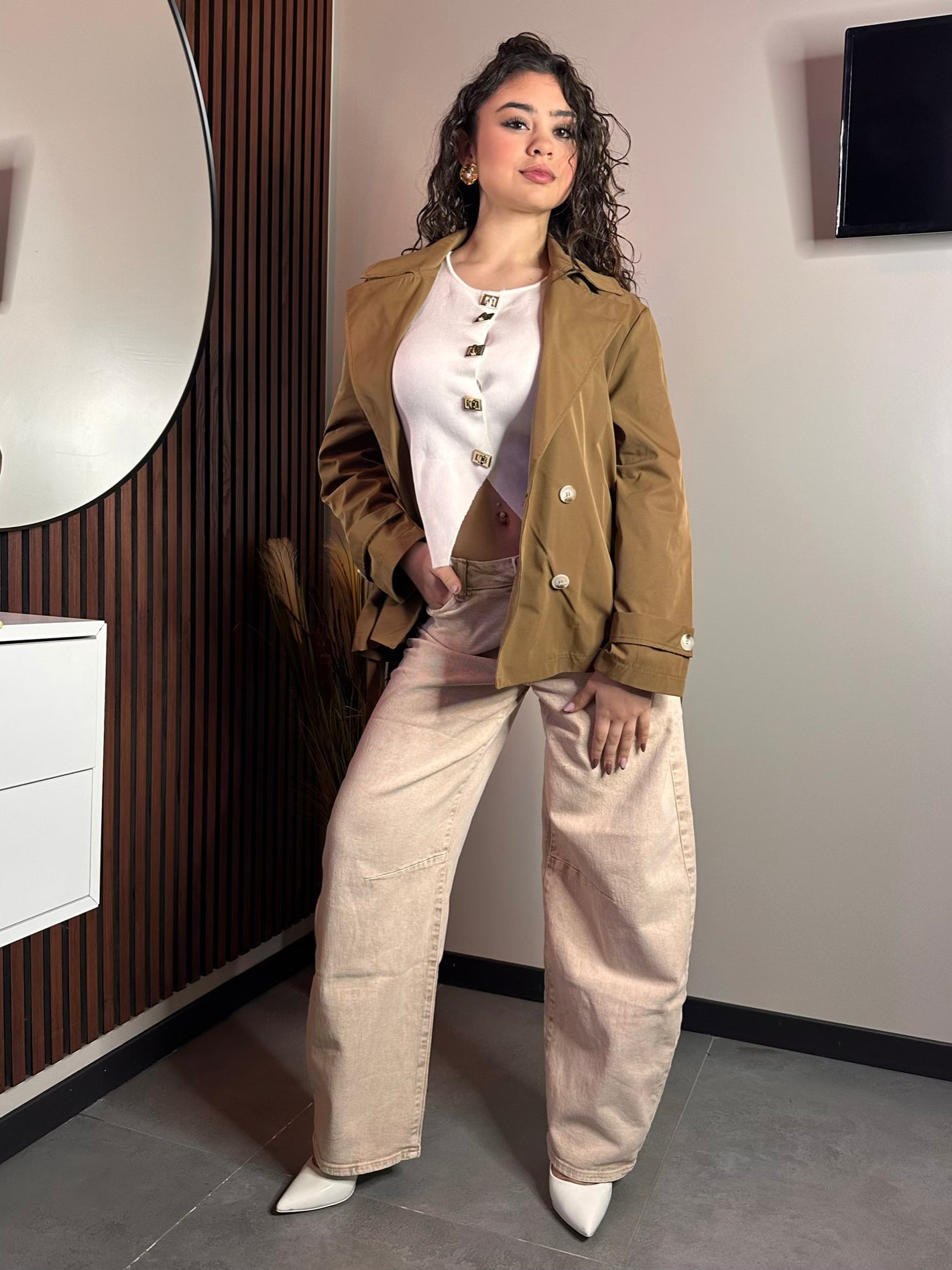 Posa a figura intera ravvicinata della modella con il trench corto color cammello indossato aperto sopra una maglia bianca e pantaloni color panna. È chiaramente visibile la parte interna della giacca e i bottoni.