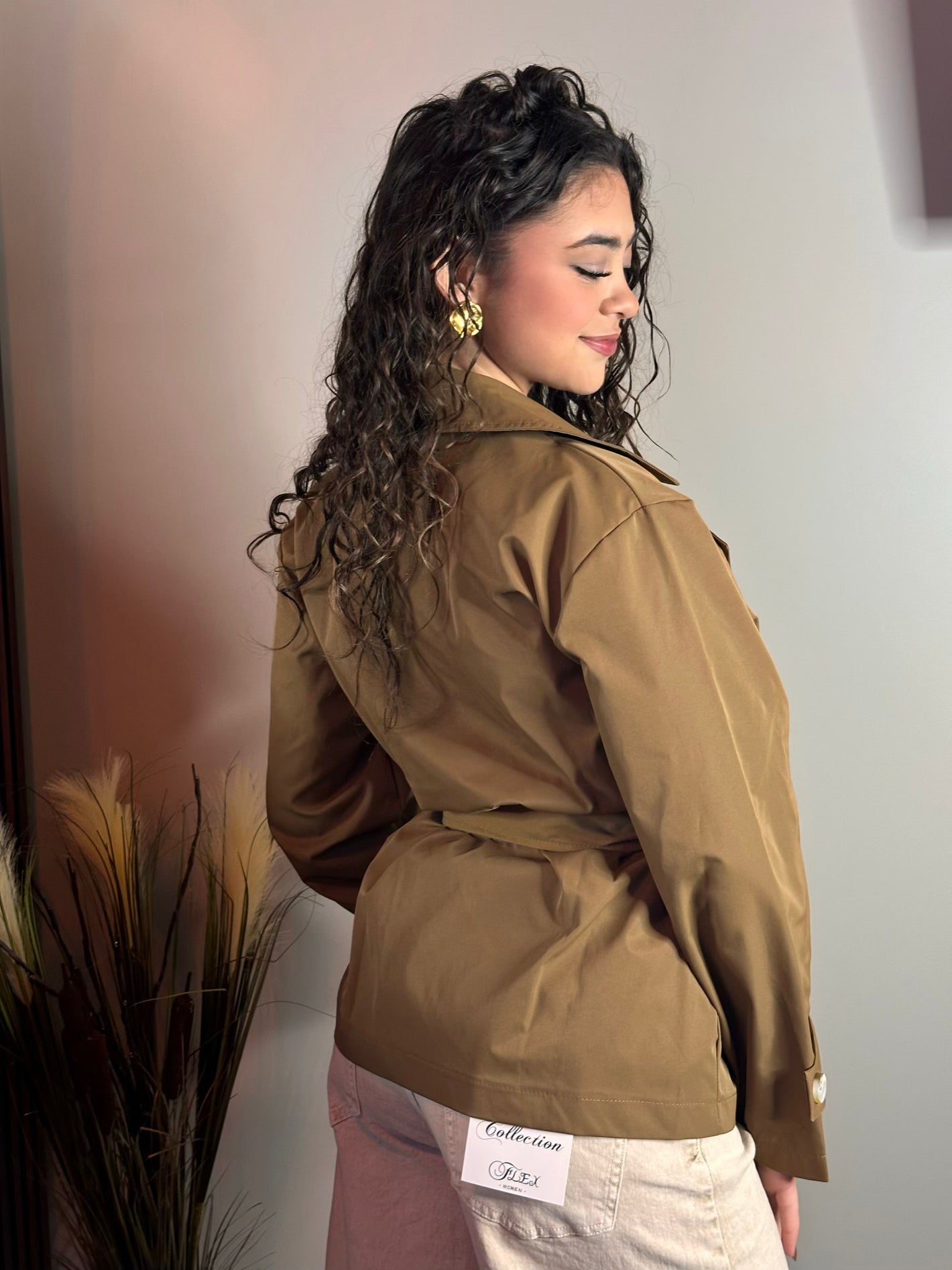 Vista posteriore della modella con il trench corto color cammello. L'immagine mostra la vestibilità del retro, la cintura che definisce il punto vita e la trama liscia del tessuto. I capelli ricci sono raccolti su un lato.